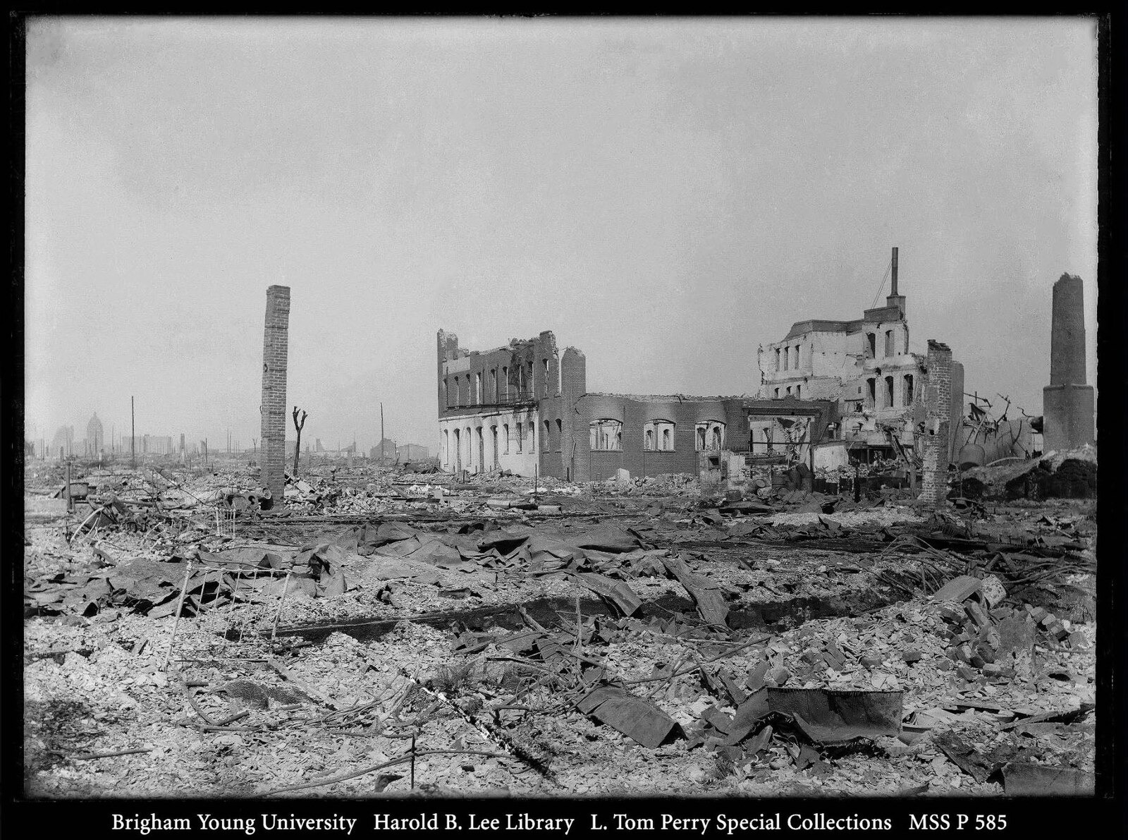 There are bed frames and some columns of bricks standing on the left and right of the photograph amidst debris and rubble from crumbled home
